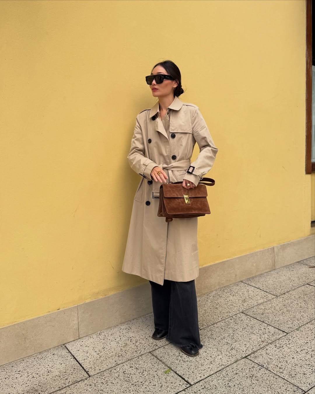 Mulher com look com calça preta e trench coat bege, combinada com bolsa marrom, encostada em parede amarela.