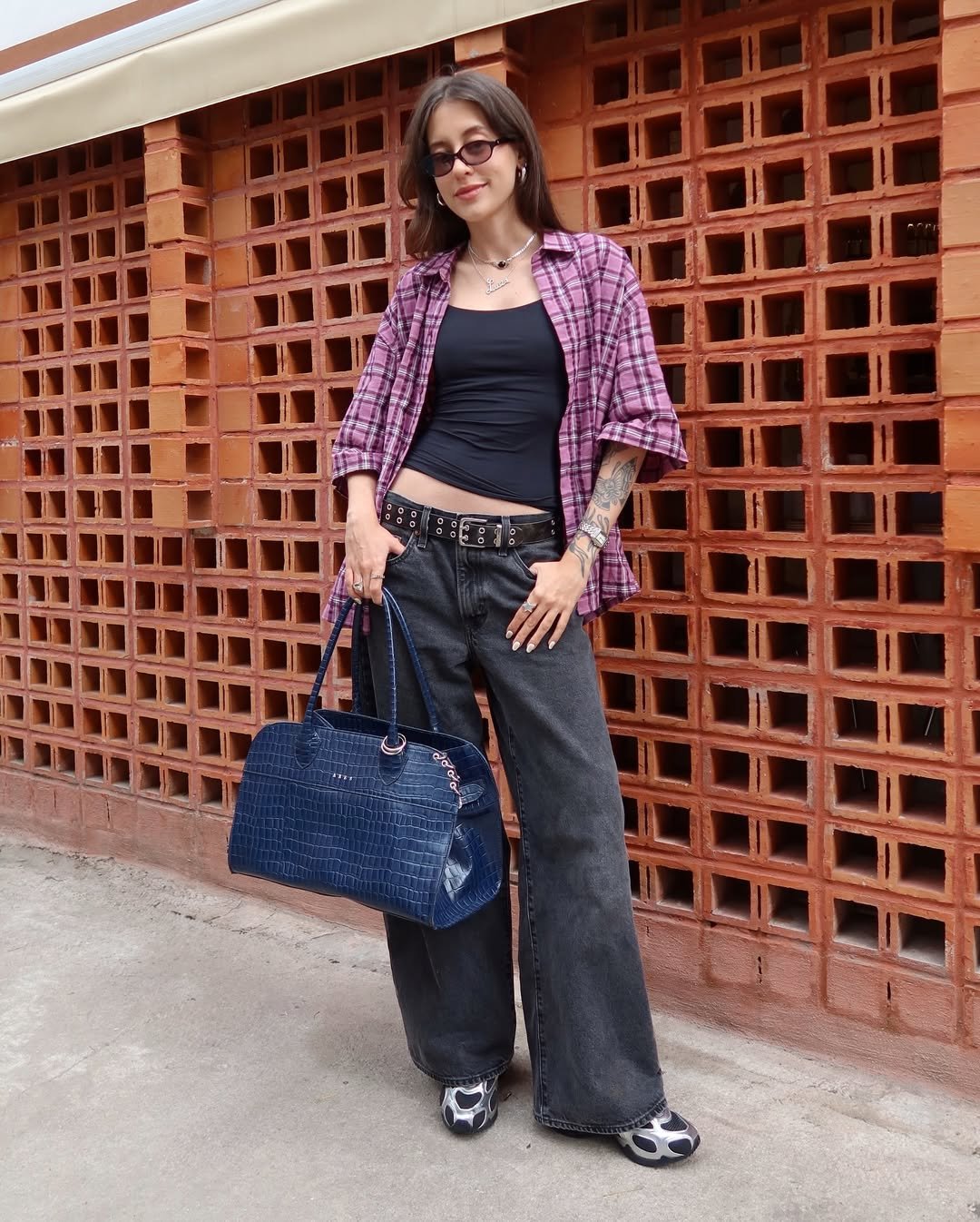 Jovem usando look com calça preta, blusa preta, camisa xadrez, cinto de tachinhas e bolsa azul.