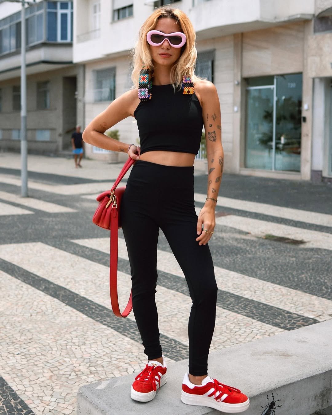 Mulher posa na calçada usando look com calça legging preta, top cropped, tênis vermelho e bolsa vermelha combinando.