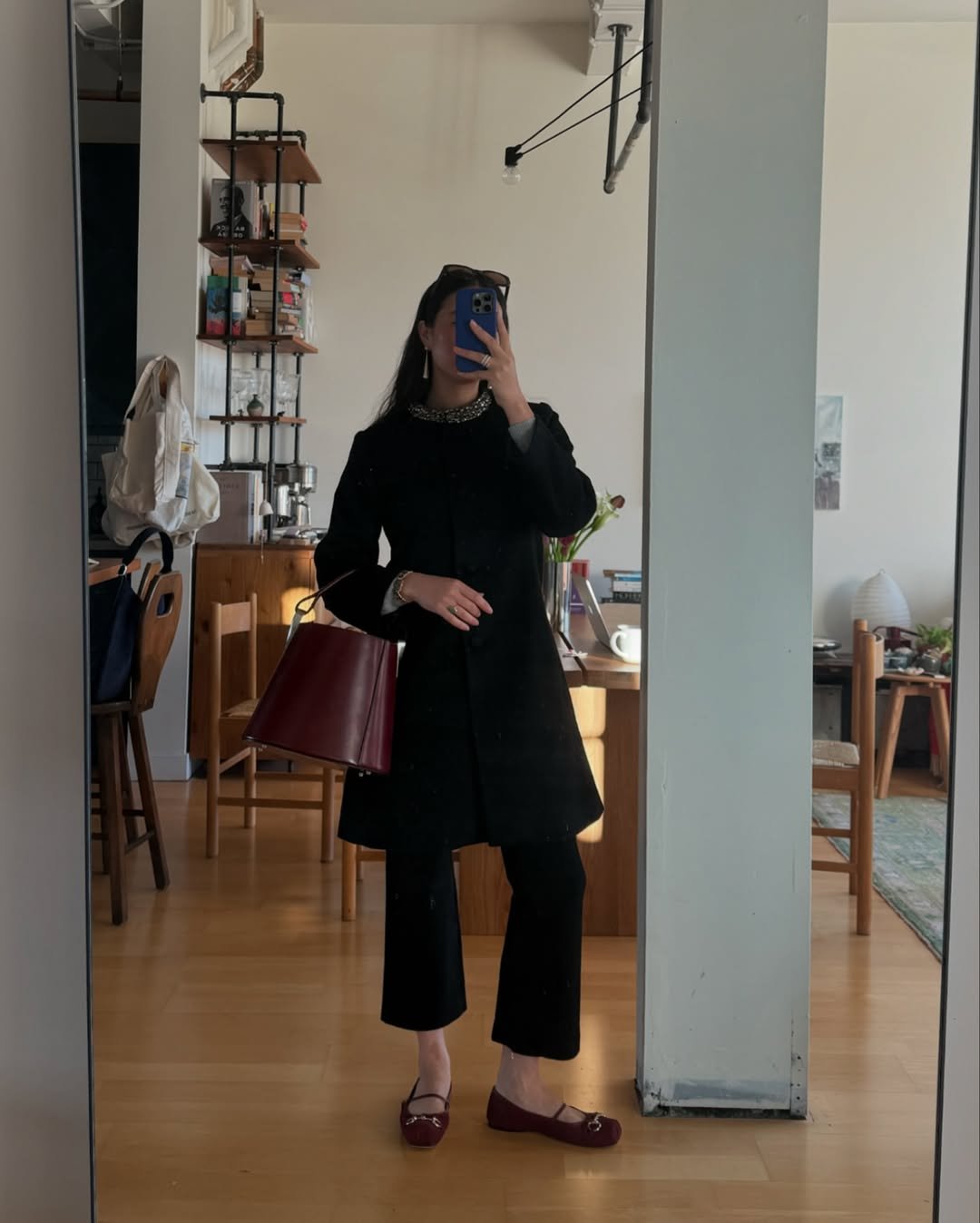 Mulher posa em frente ao espelho usando look com calça pantacourt preta, casaco longo com gola detalhada e bolsa vinho estruturada.