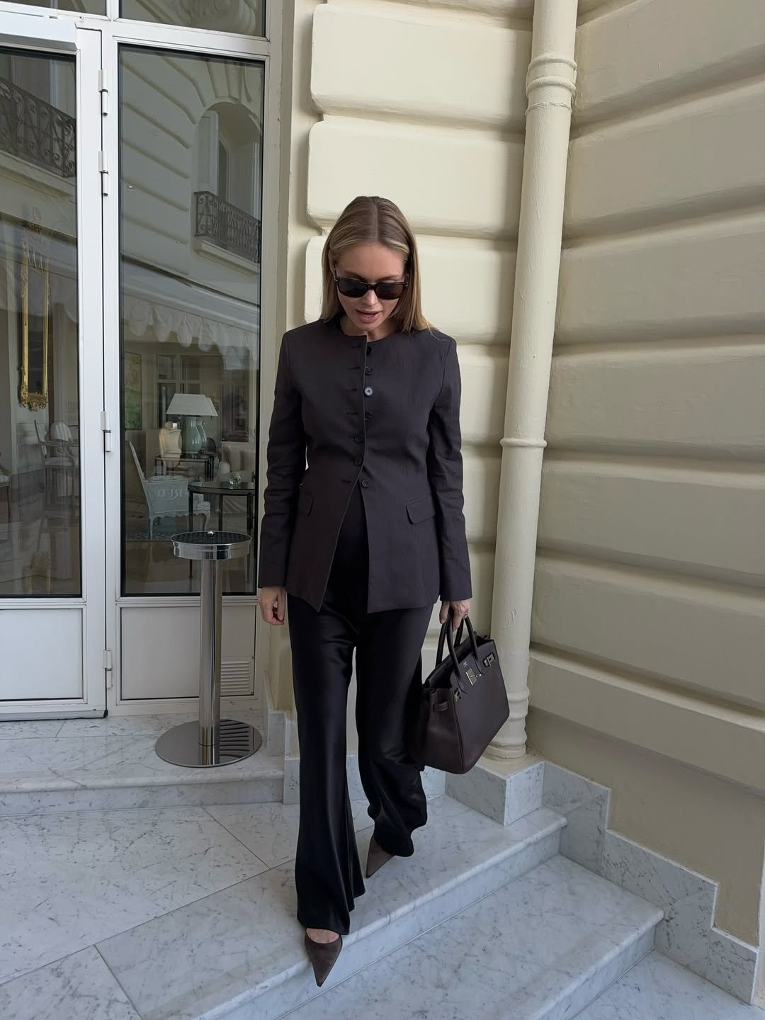 Mulher elegante desce escada usando look com calça de alfaiataria preta, blazer estruturado, salto fino e bolsa sofisticada.