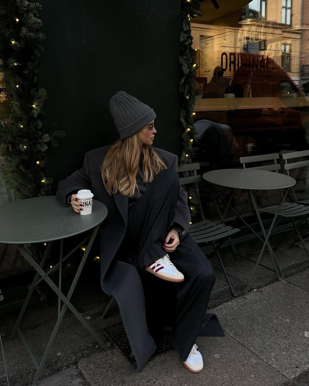 Mulher sentada em cafeteria veste look com calça preta larga, sobretudo, gorro cinza e tênis branco com listras vermelhas.