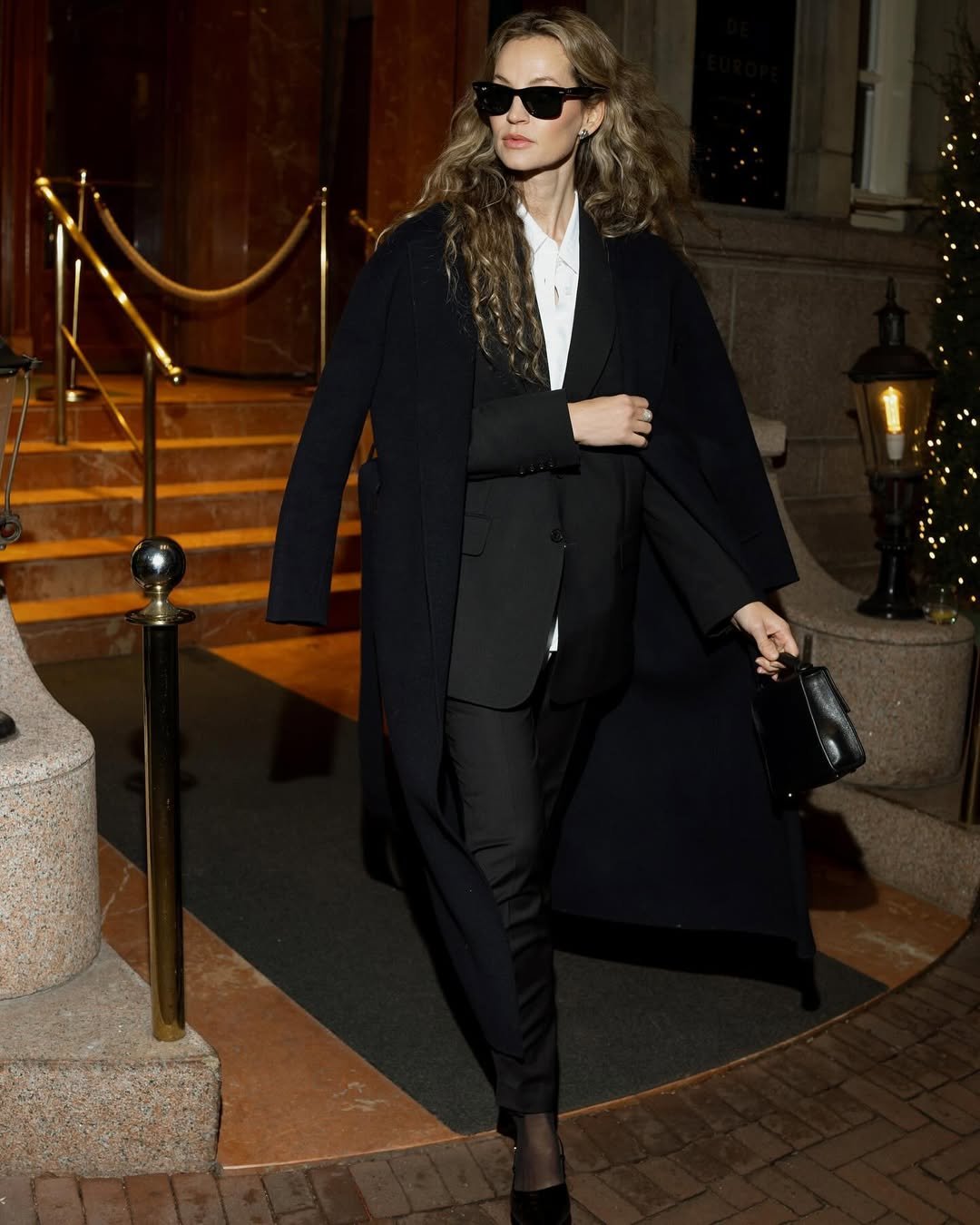 Mulher elegante sai de hotel usando look com calça social preta, camisa branca, blazer e sobretudo preto, finalizando com bolsa estruturada.