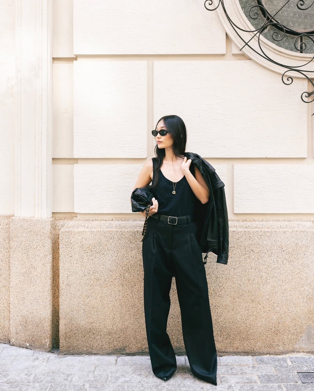 Mulher elegante usa look com calça de alfaiataria preta, regata da mesma cor, cinto largo e jaqueta de couro apoiada no ombro.