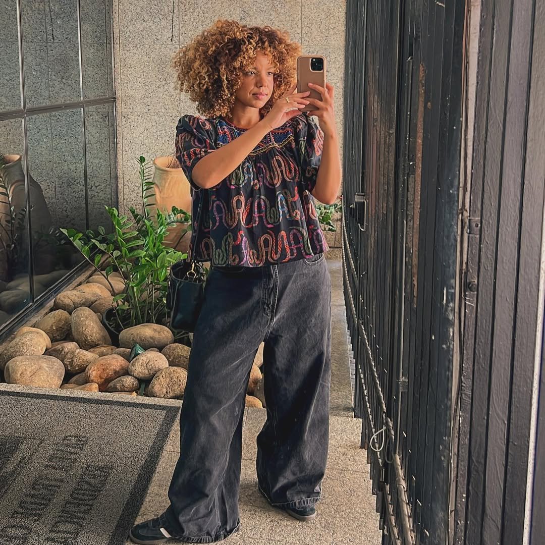 Mulher com cabelo cacheado veste blusa estampada e calça preta larga. Ensinando como montar looks com calça preta.