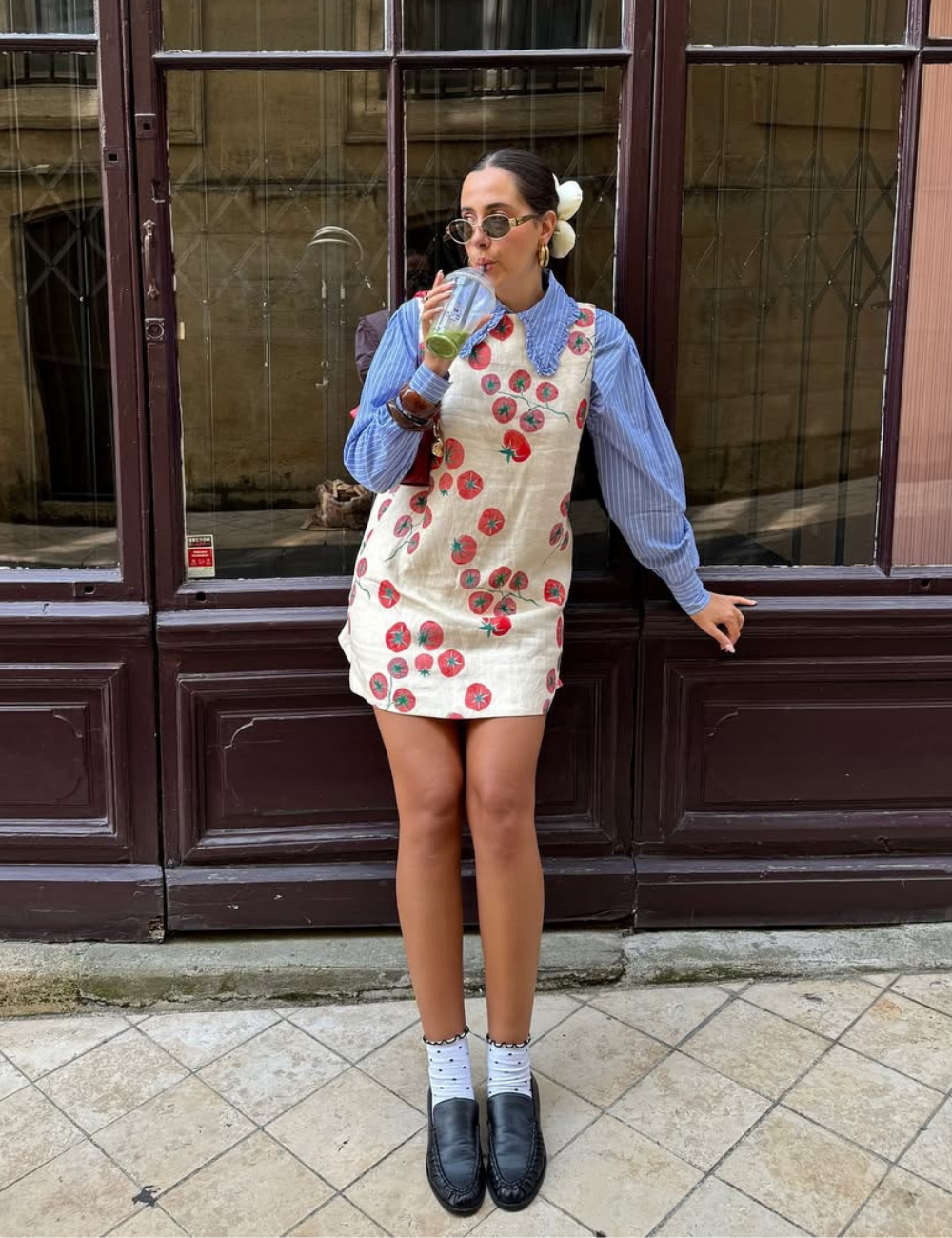 O look apresenta uma camisa listrada feminina em azul, combinada com um vestido claro decorado com estampas de tomates. O visual é complementado por óculos escuros e um coque com flor, trazendo um toque de frescor.