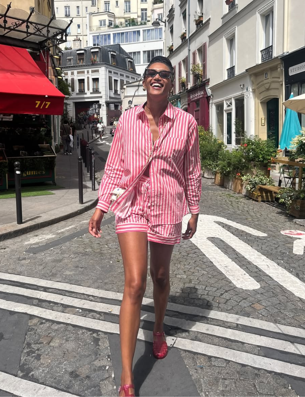 Look com camisa listrada feminina destaca uma combinação de estilo e conforto. A modelo veste uma camisa de listras rosa e branca, que traz um toque de frescor e elegância. A peça é complementada por shorts, criando um visual casual e moderno.