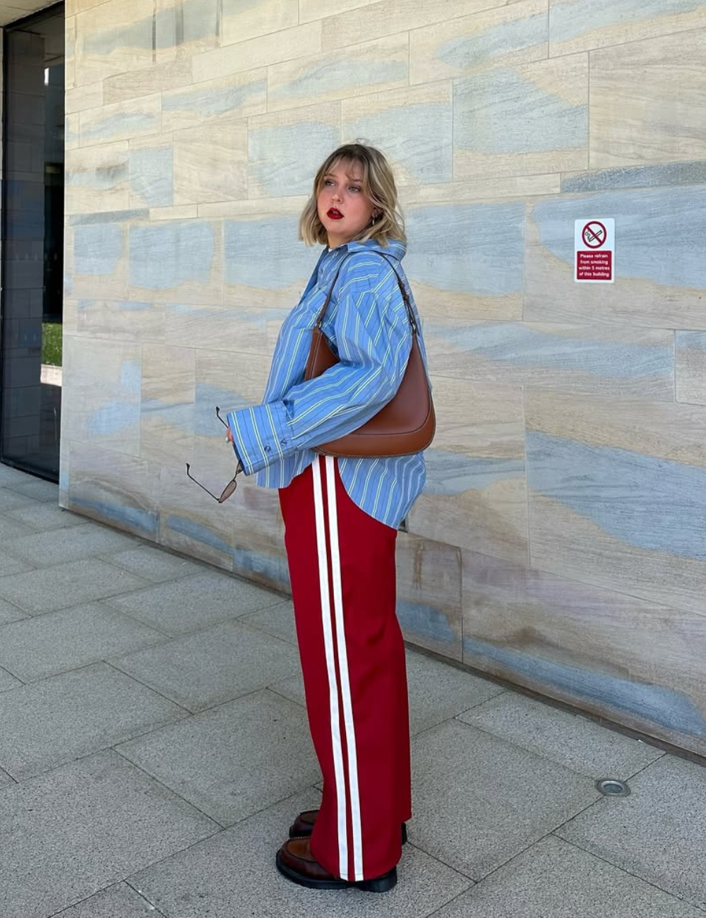 Look com camisa listrada feminina destaca uma blusa oversized em tons de azul com listras verticais, combinada com calças vermelhas que possuem listras brancas laterais. A modelo, com cabelo curto e ondulado, usa batom vermelho.