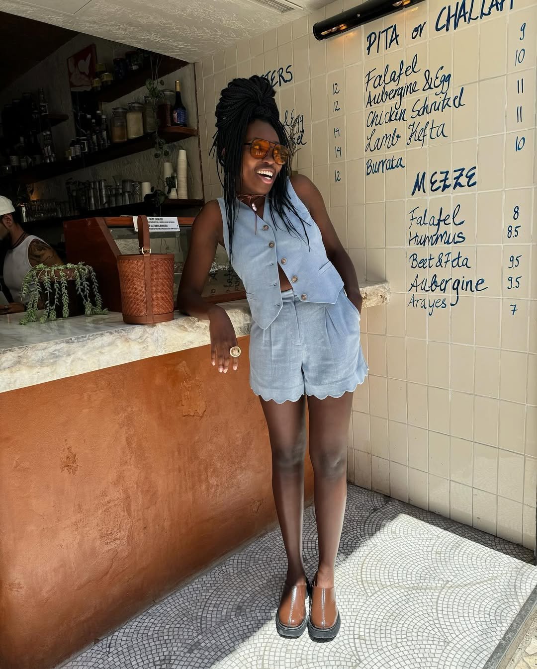 Pessoa sorridente com look com colete azul claro e shorts combinando, em pé próximo a um balcão em restaurante.