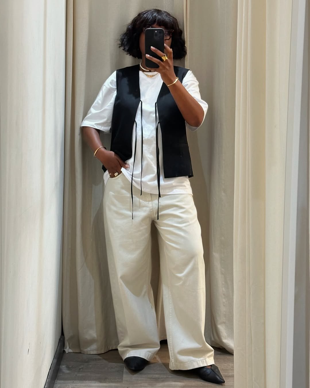 Pessoa em provador usando camiseta branca, look com colete preto e calça larga, segurando celular.