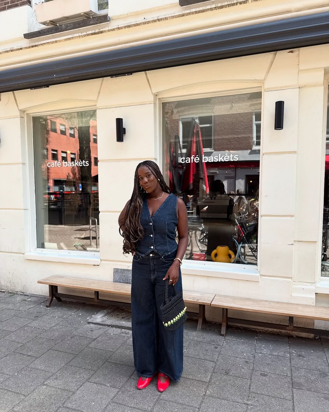 Pessoa em frente a um café, usando look com colete jeans e calça larga, segurando uma bolsa preta.