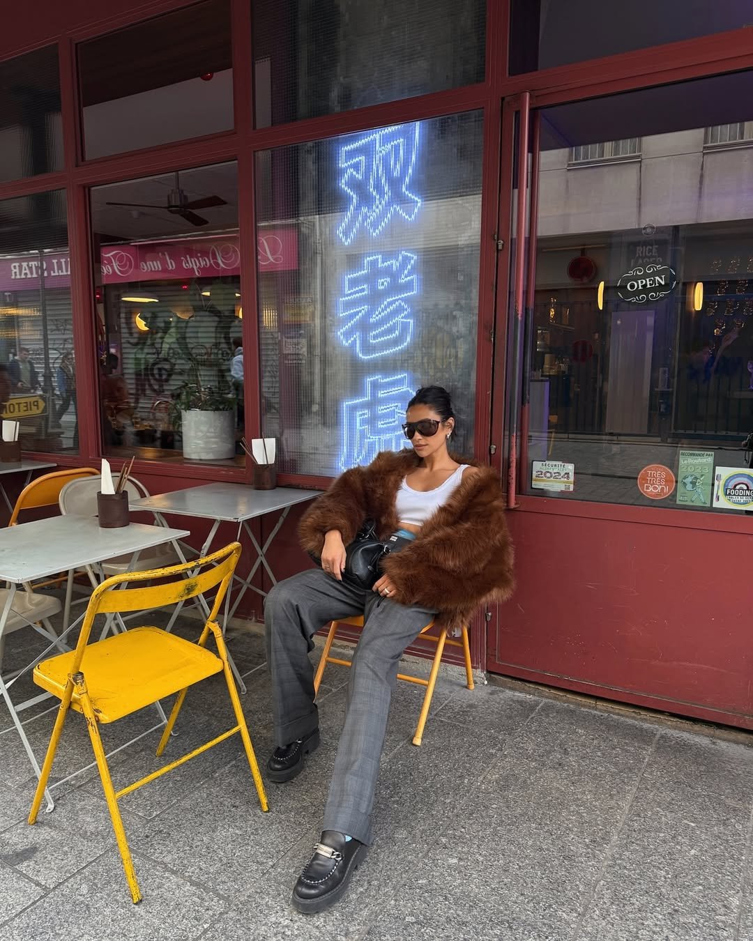 Mulher com look com mocassim tratorado, calça de alfaiataria cinza, top branco, casaco de pelo marrom e bolsa preta.