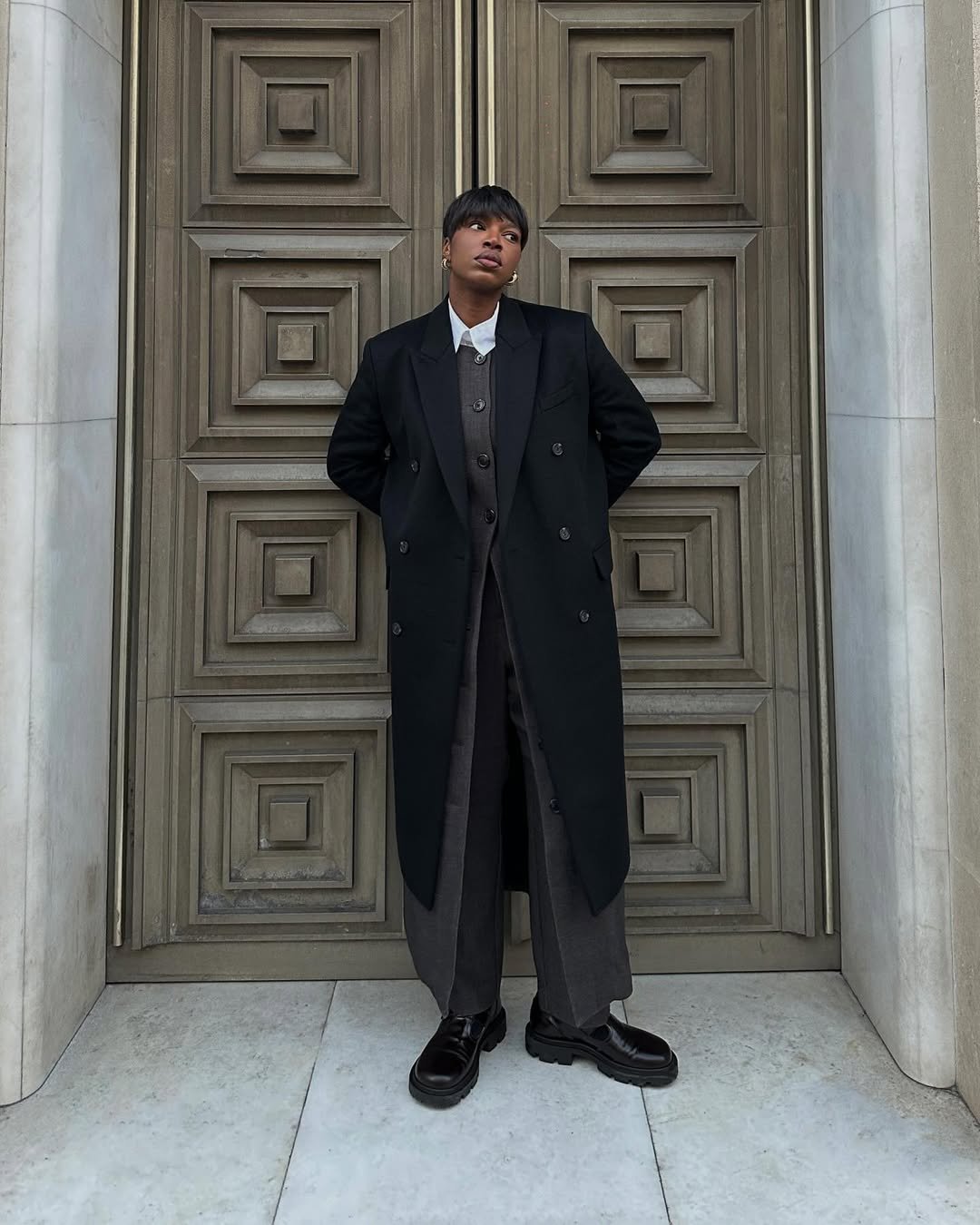 Mulher com look com mocassim tratorado, casaco longo preto, conjunto de alfaiataria cinza e camisa branca, posando em frente a uma porta.