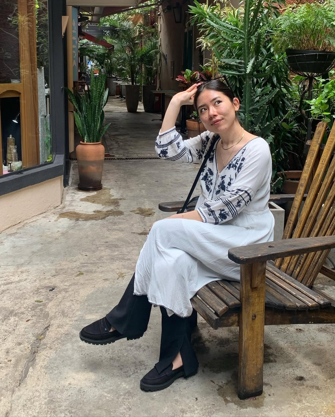 Mulher com look com mocassim tratorado, vestido branco com detalhes bordados e calça preta, sentada em banco de madeira.