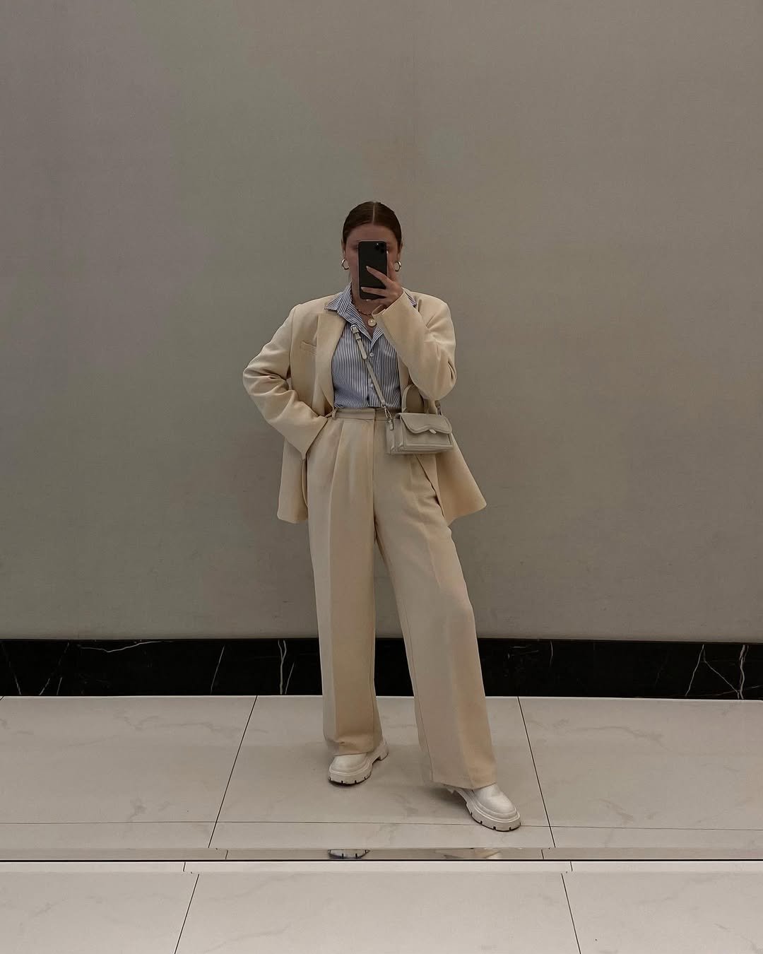 Mulher com look com mocassim tratorado off white, conjunto de alfaiataria bege, camisa listrada e bolsa pequena.