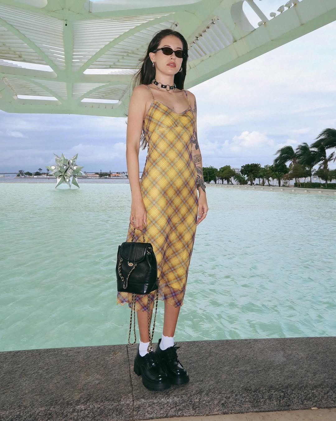 Mulher com look com mocassim tratorado, vestido midi xadrez amarelo, óculos escuros, meias brancas e bolsa preta.