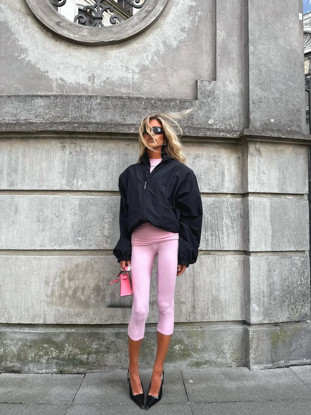 Mulher look rosa bebê com calça capri, jaqueta preta e bolsa rosa segura, em frente a parede de pedra.