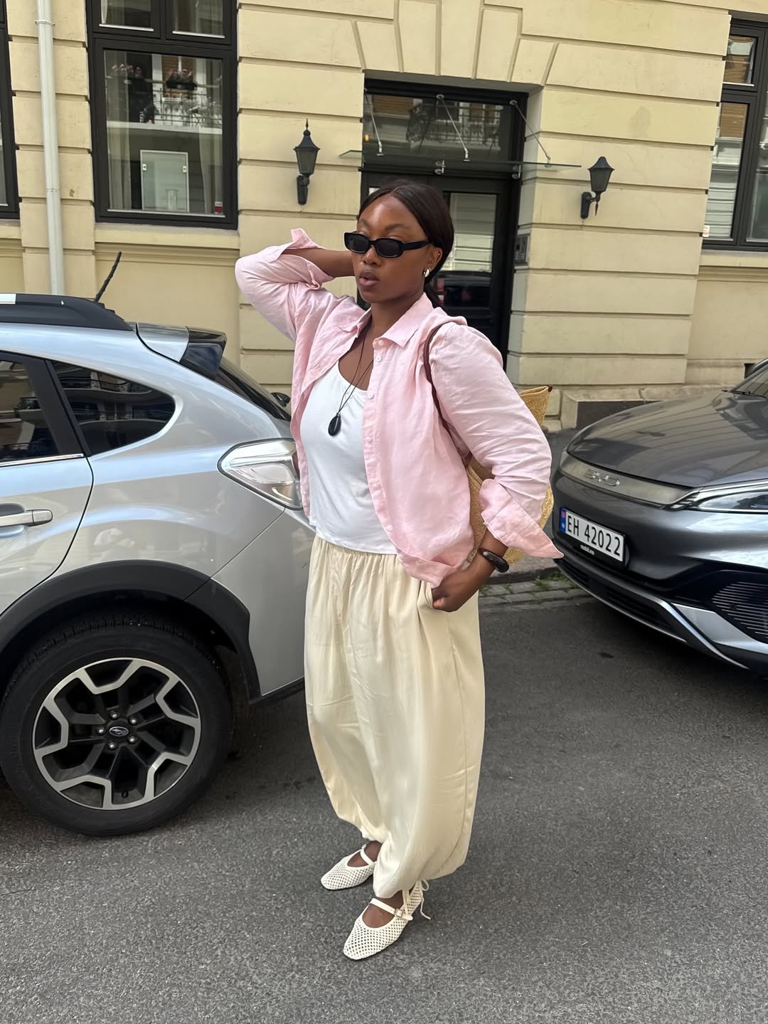 Mulher com look com camisa rosa bebê, óculos escuros e calça bege, posando entre dois carros.