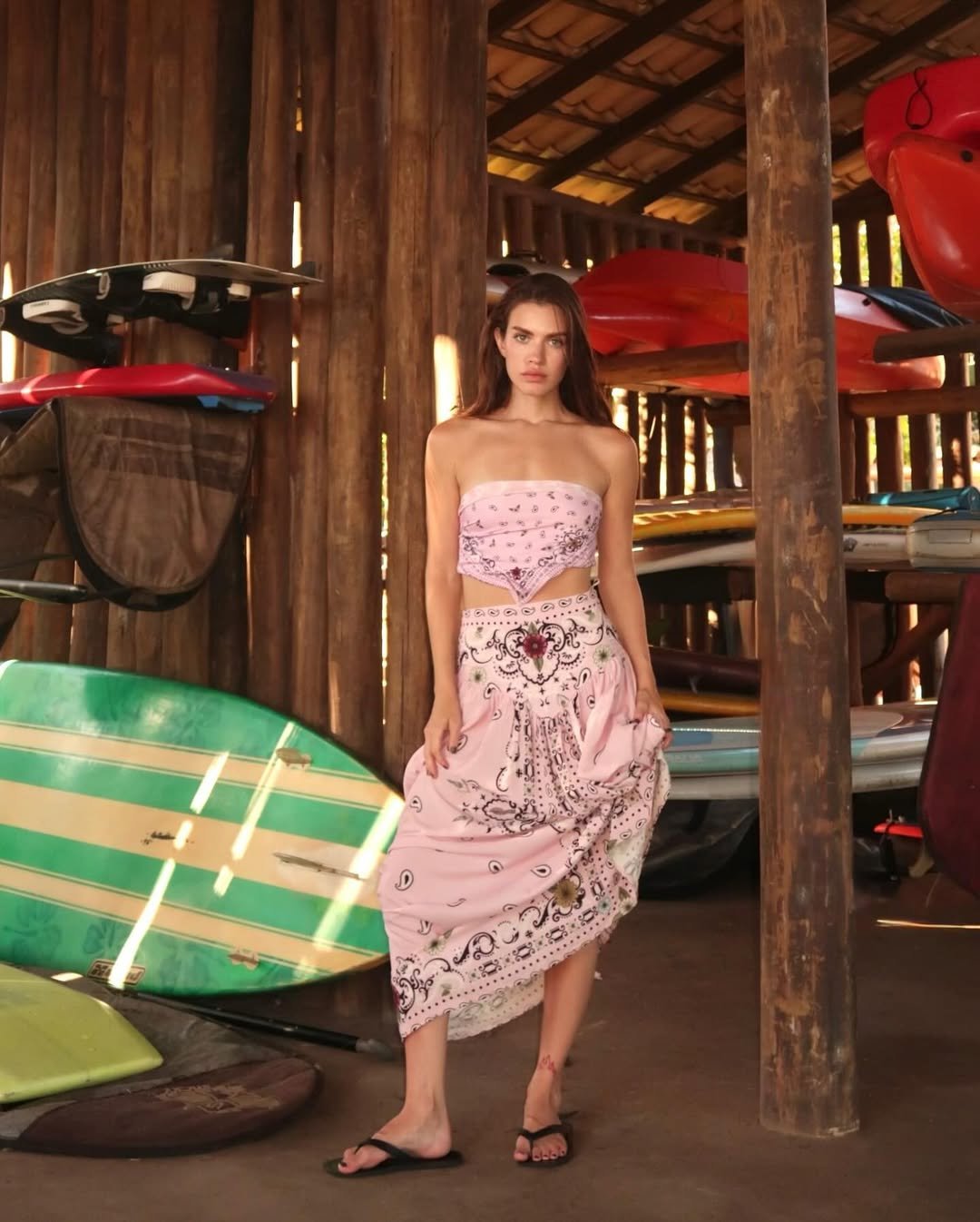 Mulher com roupas rosa bebê em um galpão com pranchas de surfe atrás.