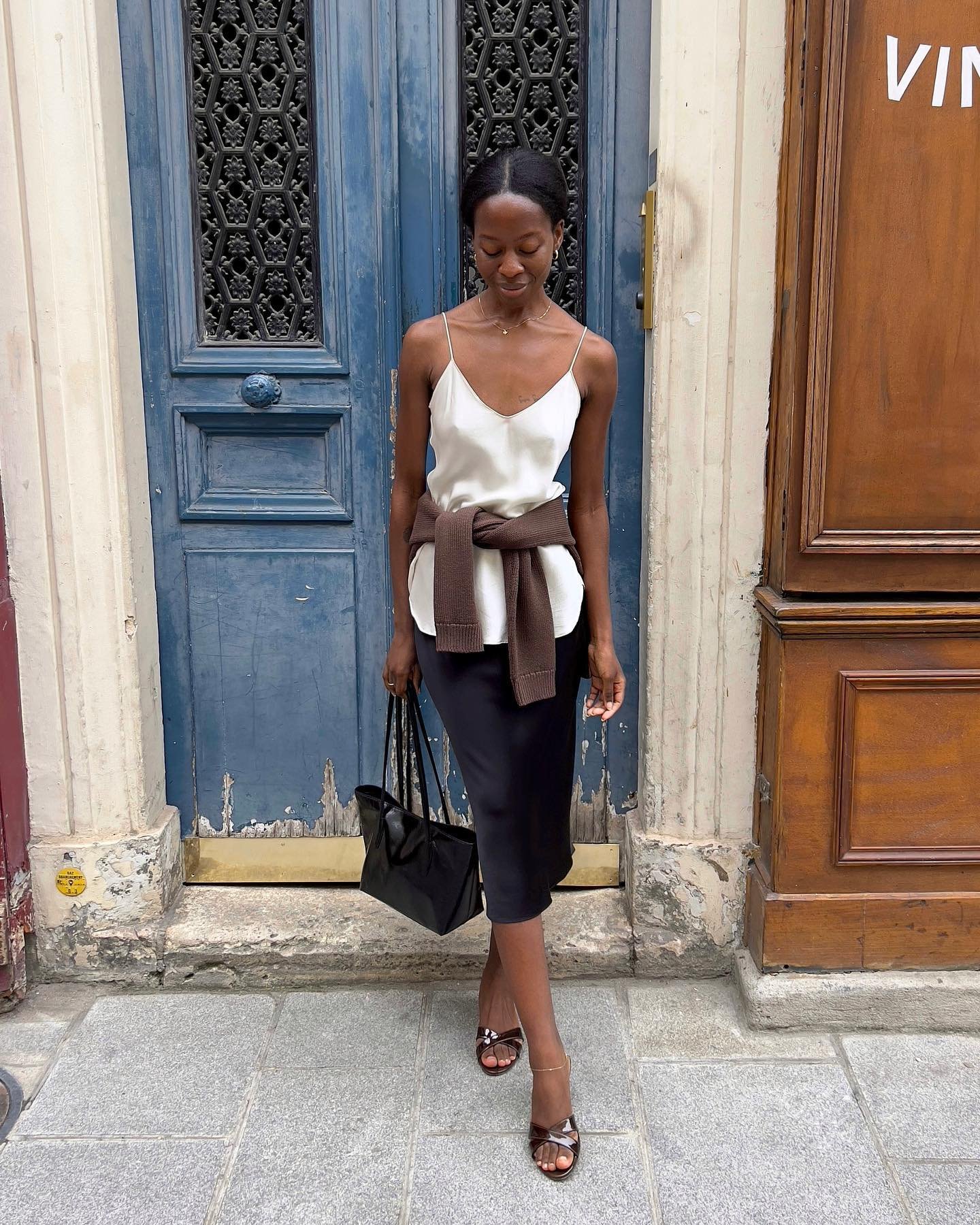 Uma pessoa com look com saia de cetim preta, blusa clara e suéter marrom em frente a uma porta azul.