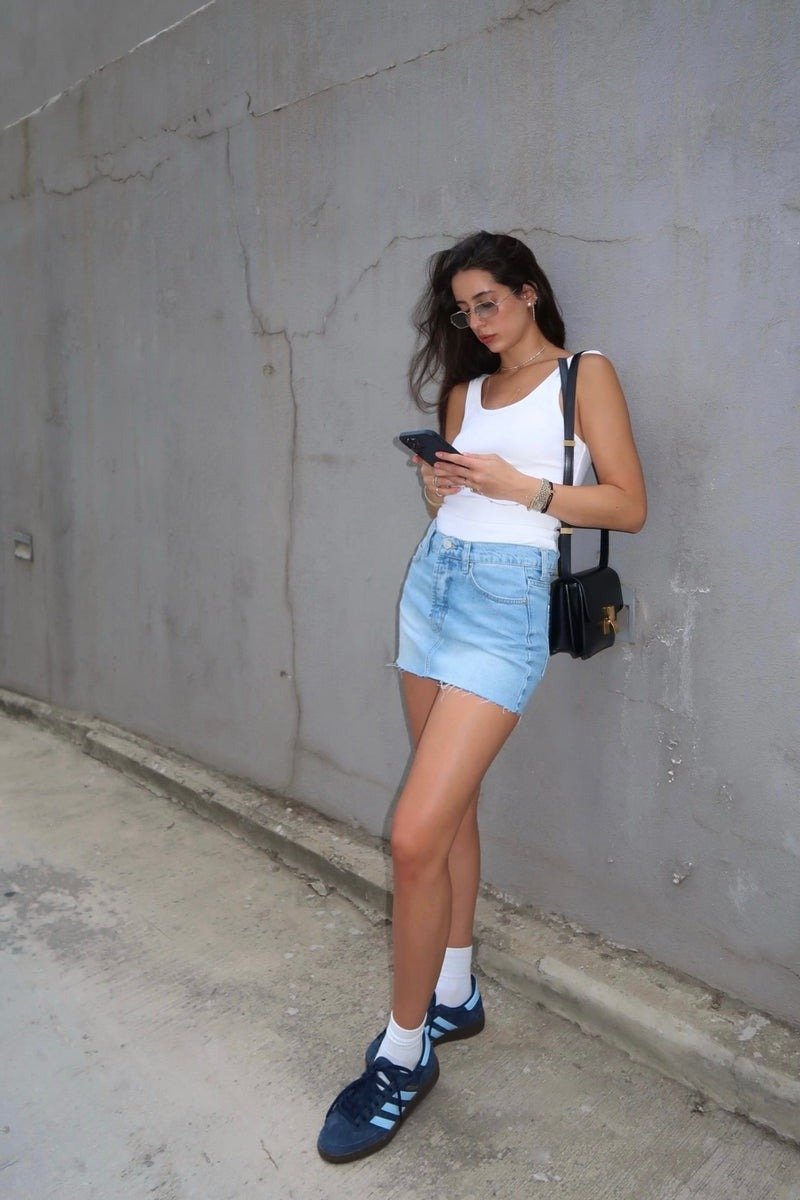 Look com saia jeans curta apresenta uma combinação casual e moderna. A saia, de lavagem clara e com corte reto, destaca as pernas, enquanto a blusa branca de alças finas traz leveza ao visual. O uso de tênis escuros com detalhes em azul e meias brancas ad