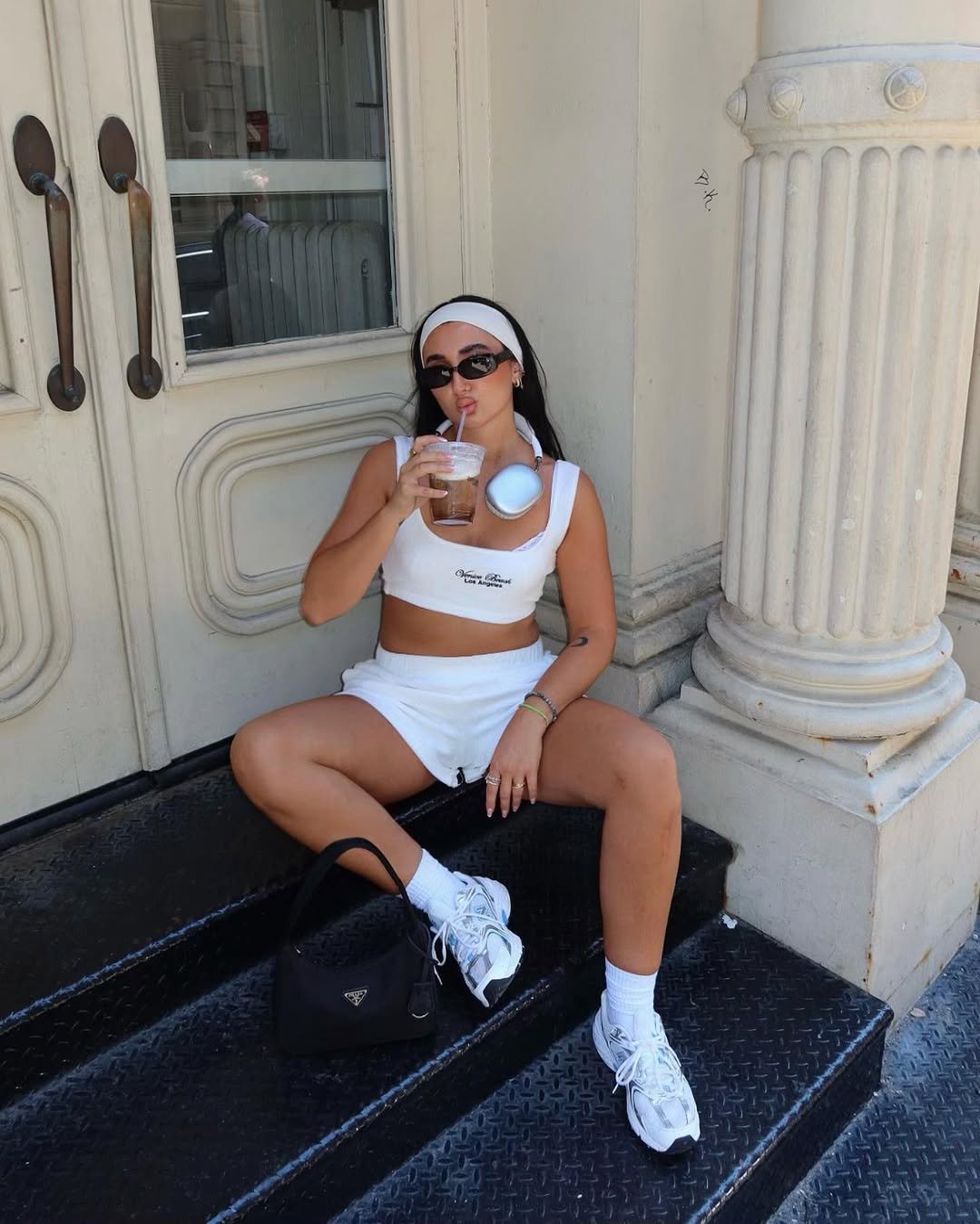 Pessoa sentada nas escadas, usando look de academia roupa esportiva casual, óculos de sol e tomando uma bebida gelada.