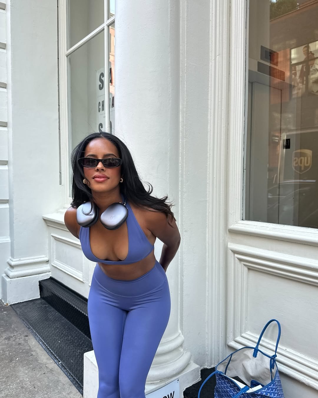 Mulher em look de academia azul, com fones no pescoço e óculos escuros, em frente a prédio urbano.