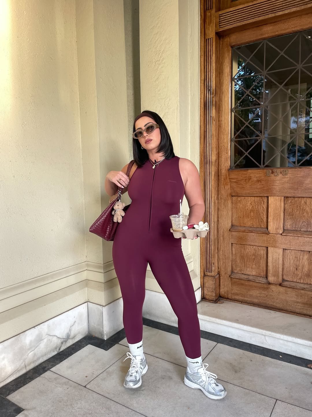 Mulher com look de academia roxa, tênis brancos, óculos escuros e bolsa, segura bebidas em local elegante.