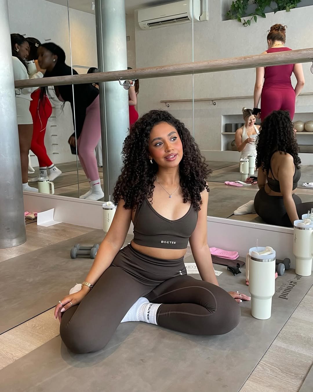 Pessoa sentada no chão de academia, usando conjunto esportivo marrom, cercada por acessórios de treino. Look de academia.