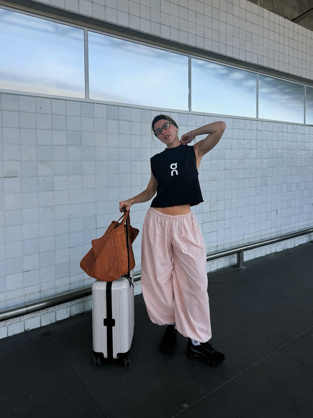 Mulher usando look com calça rosa bebê e blusa preta ao lado de mala e bolsa. Ambiente urbano com parede de azulejos.