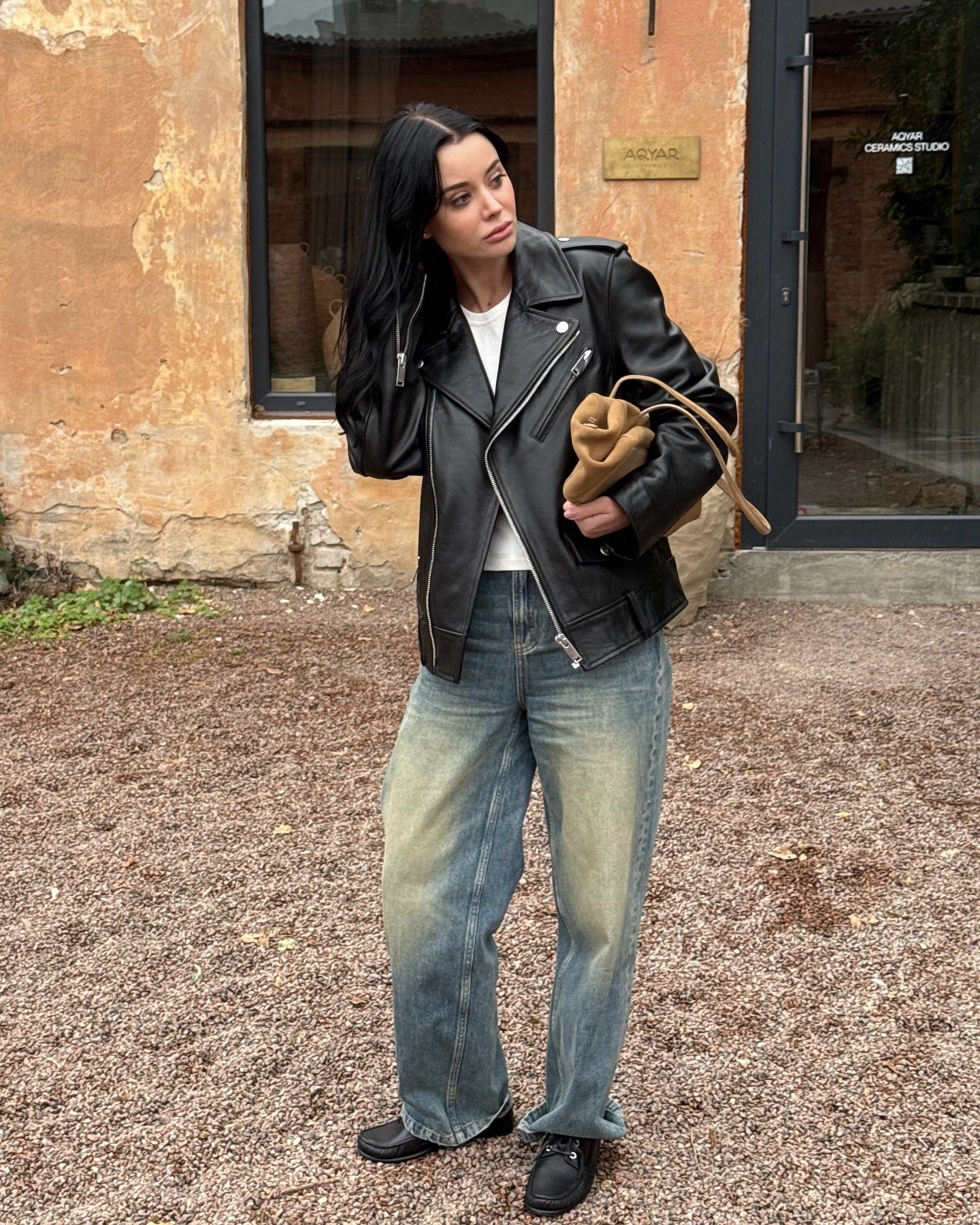 O look meia-estação apresenta uma combinação estilosa de jaqueta de couro preta e jeans oversized. A jaqueta, com detalhes em zíper, confere um ar contemporâneo, enquanto a camiseta branca traz leveza.