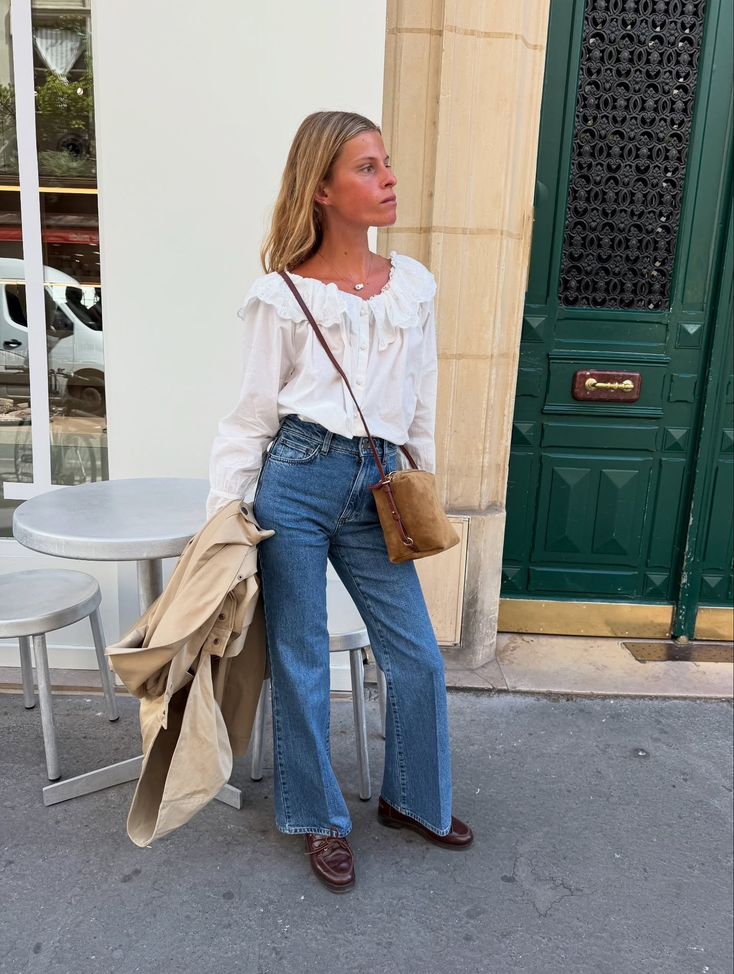 O look meia-estação apresenta uma combinação elegante e descontraída. A blusa branca com detalhes em babados traz leveza, enquanto a calça jeans ampla oferece conforto e estilo. O casaco bege, pendurado no braço, é perfeito para temperaturas amenas.