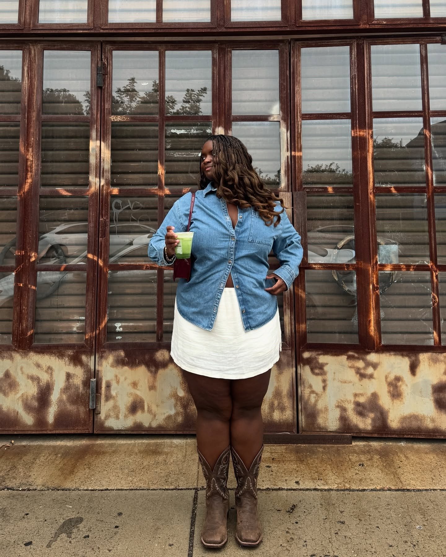 O look meia-estação apresenta uma mulher com cabelo cacheado e trançado, vestindo uma camisa jeans oversized, que complementa uma saia branca. Ela usa botas estilo cowboy e segura uma bebida verde, transmitindo um ar descontraído e estiloso.