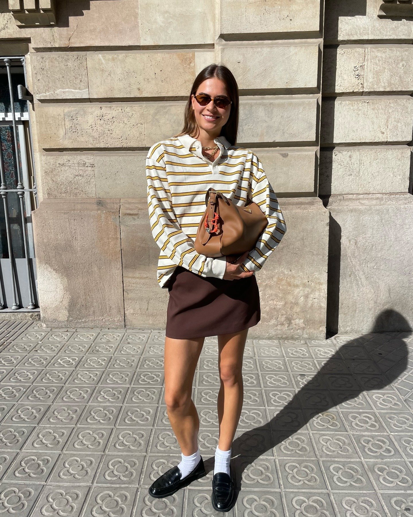 O look meia-estação apresenta uma combinação estilosa de camisa listrada em tons de amarelo e branco, com uma saia marrom curta. O uso de meias brancas e loafers pretos confere um toque contemporâneo.