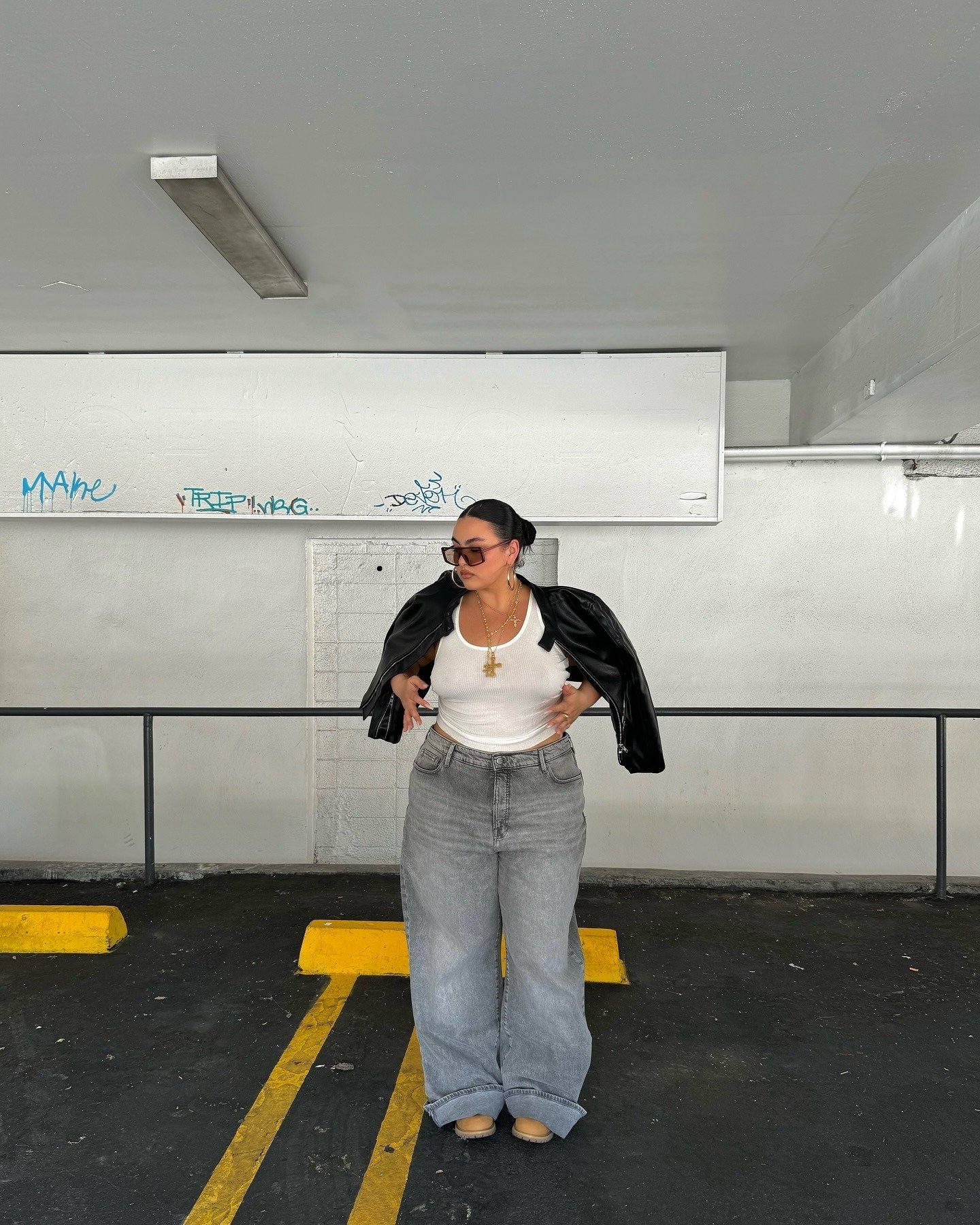 O look meia-estação apresenta uma combinação estilosa e descontraída. A modelo usa uma regata branca, complementada por um blazer de couro preto que adiciona sofisticação ao visual. As calças jeans largas em tom cinza oferecem conforto e modernidade.