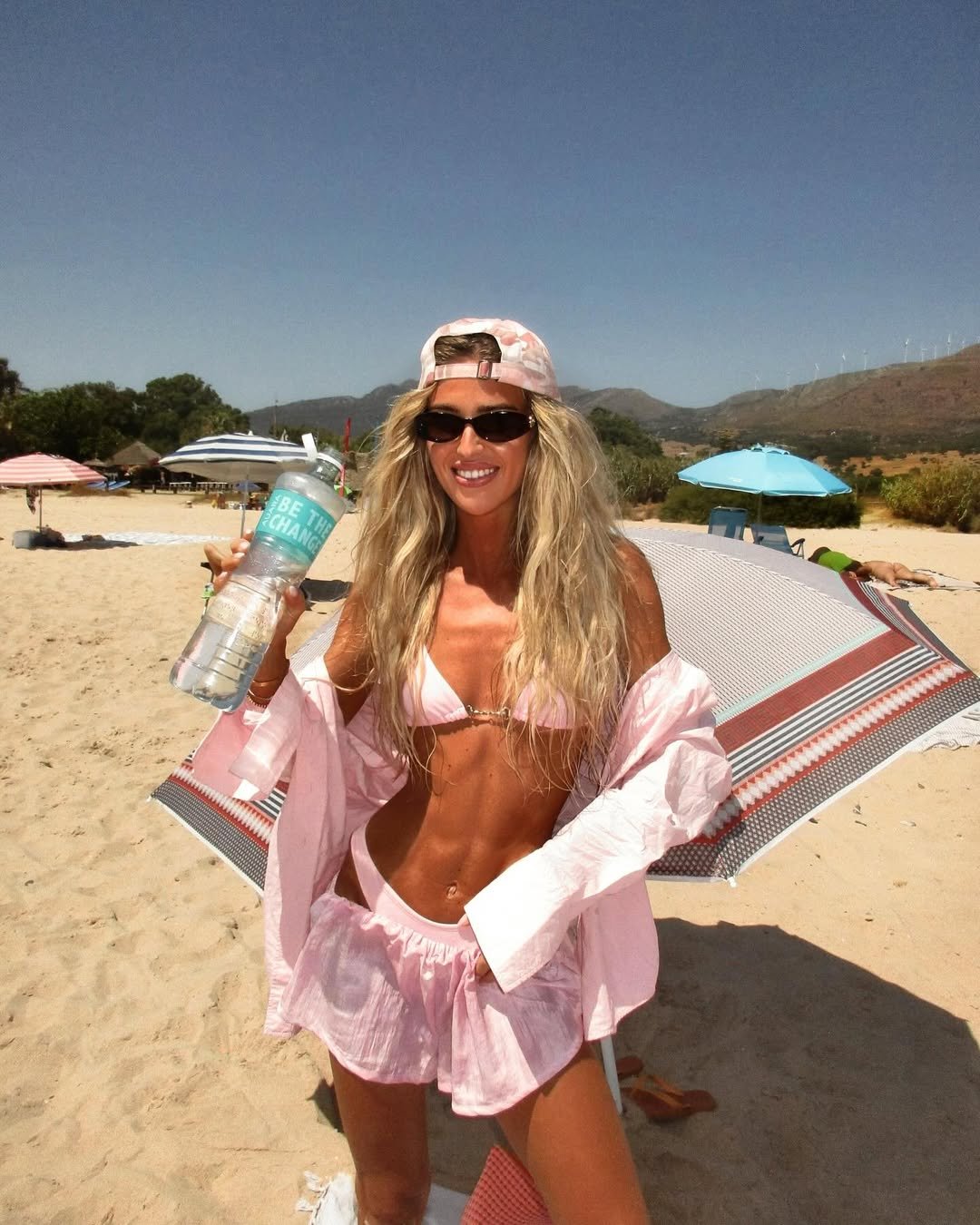 Mulher com look de praia rosa bebê, camiseta aberta, boné e óculos escuros, segurando garrafa d'água.