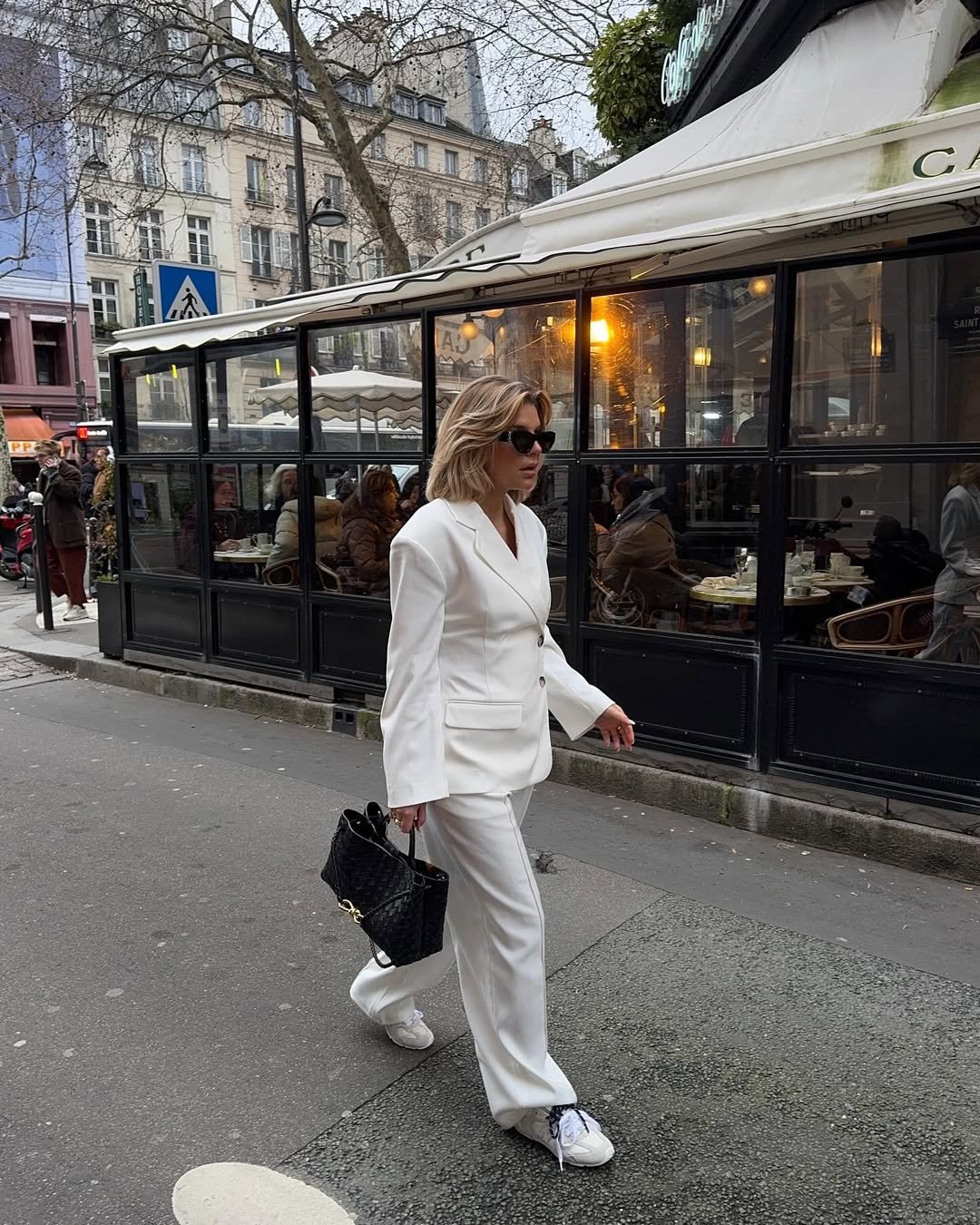 Pessoa com terno branco, tênis branco e óculos de sol caminha em frente a café na cidade. Look com tênis branco