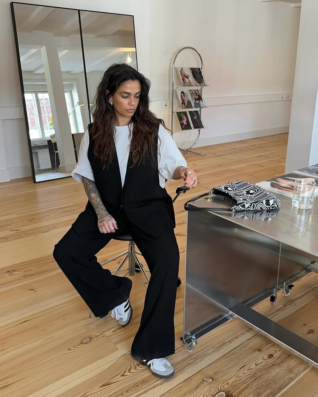 Ela está sentada em um banco, usando um look moderno com terno preto e tênis branco.