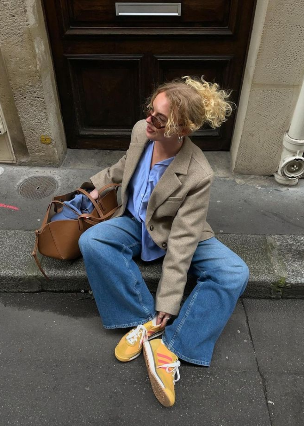 A adidas SL 72 é destacada nos pés de uma mulher que exibe um estilo moderno e casual. Ela usa um blazer oversized em tom neutro, combinado com uma blusa azul clara e jeans largos, criando um look equilibrado. Os tênis amarelos com detalhes em vermelho e