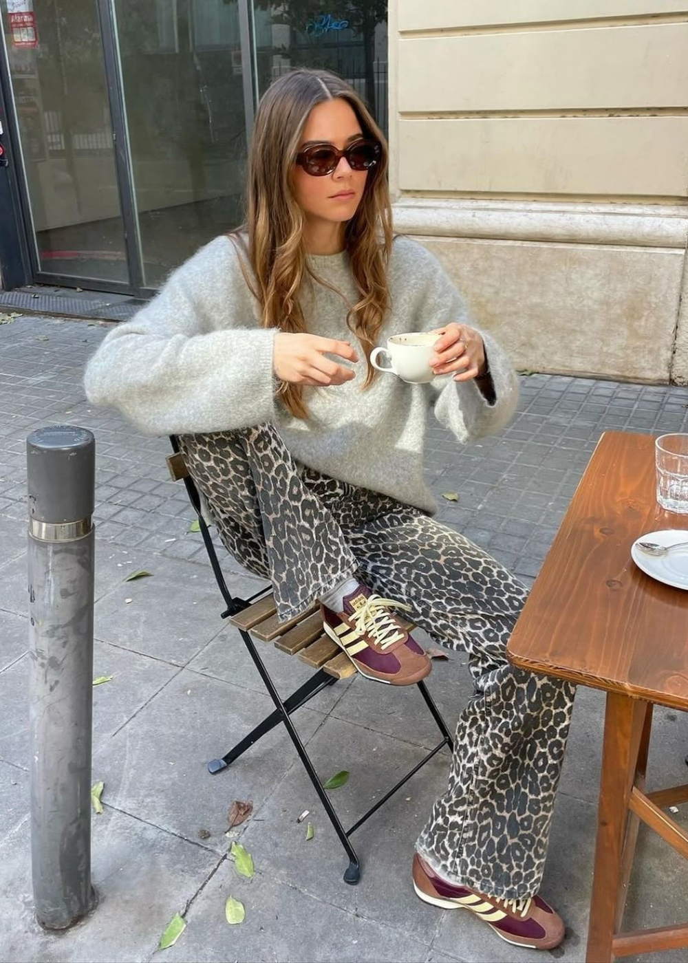 A imagem apresenta uma mulher sentada em uma cadeira, usando um suéter cinza e calças com estampa de leopardo, que trazem um toque ousado ao visual. Nos pés, destaca-se o modelo adidas SL 72, em tons de marrom e detalhes em amarelo.