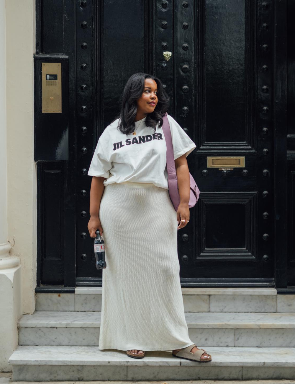 Mulher com camiseta branca, saia longa e sandálias estilo papete em frente a porta preta.