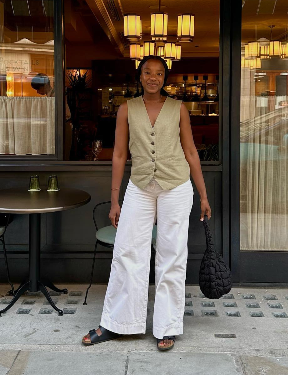 Pessoa em frente a um restaurante usa colete bege, calça branca larga e papete preta, segurando bolsa preta, destacando looks bonitos.