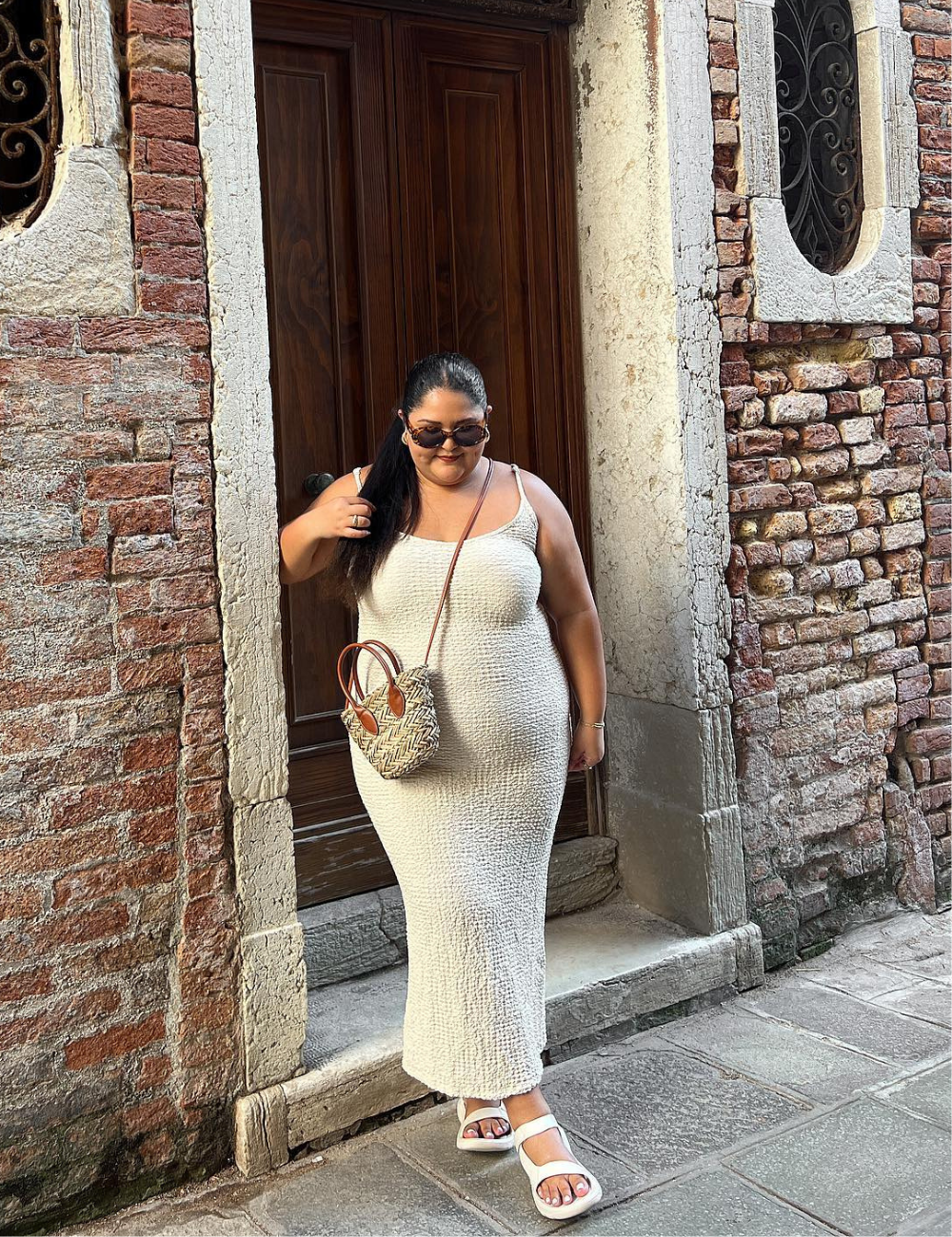 Pessoa usando vestido bege, sandálias papete brancas, óculos de sol e bolsa de palha. Fundo com parede de tijolos.