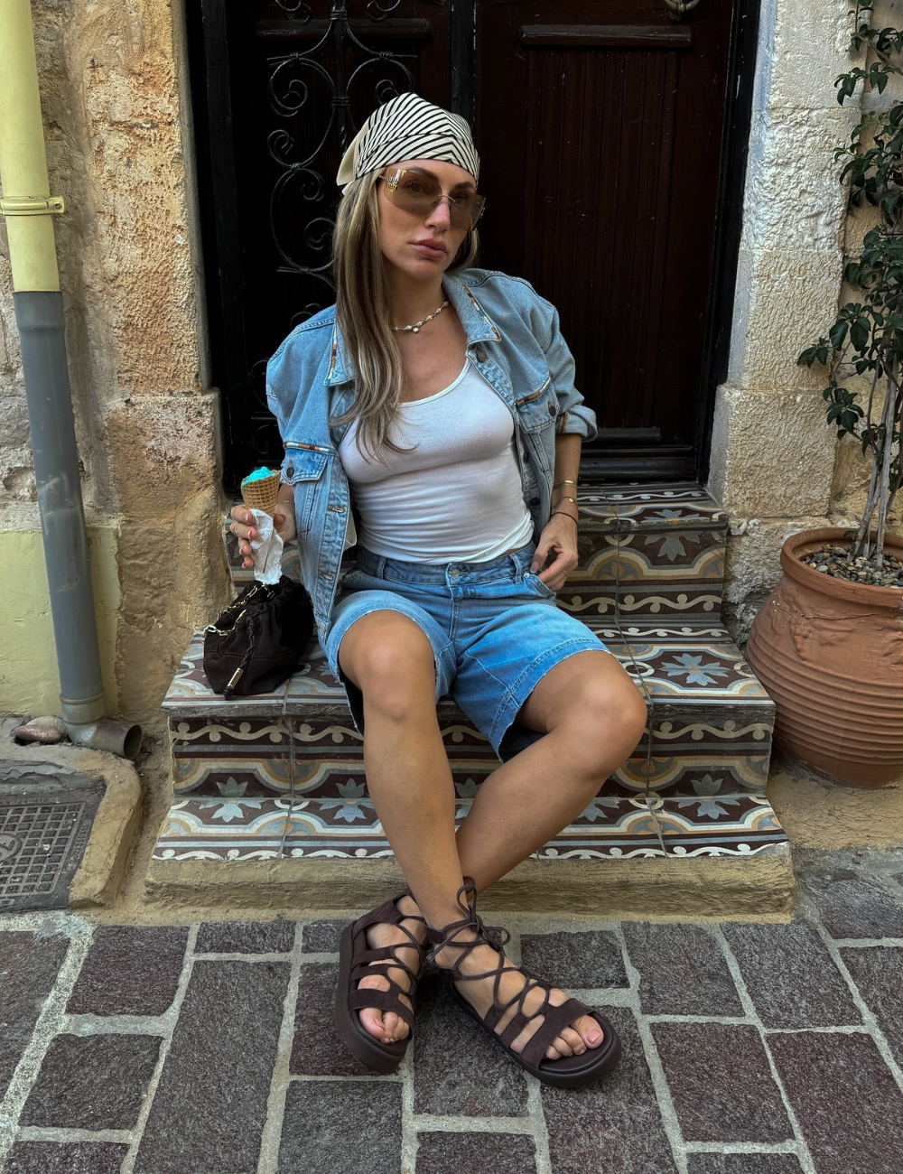 Pessoa com bandana, jaqueta jeans e papete, sentada em degrau decorado, tomando sorvete.