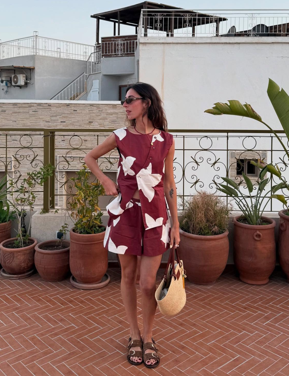 Mulher usa conjunto vermelho com estampas brancas, chinelos estilosos e bolsa de palha, em terraço com plantas. Um dos looks bonitos com papete.