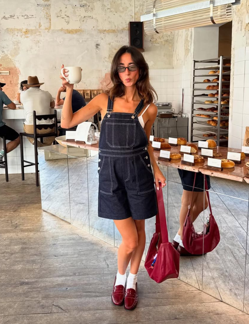 O look apresenta um macacão curto em jeans escuro, ideal para um visual casual e estiloso. A combinação com uma blusa branca e sapatos oxford burgundy traz um toque moderno. Acessórios como óculos escuros e uma bolsa transversal complementam a produção.