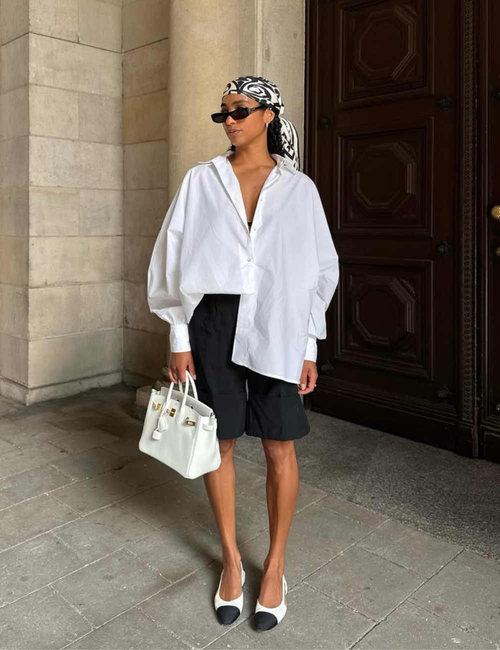 O look apresenta uma camisa branca oversized, combinada com shorts pretos, criando um contraste elegante e descontraído. A modelo usa um lenço estilizado na cabeça e óculos escuros, adicionando um toque moderno.