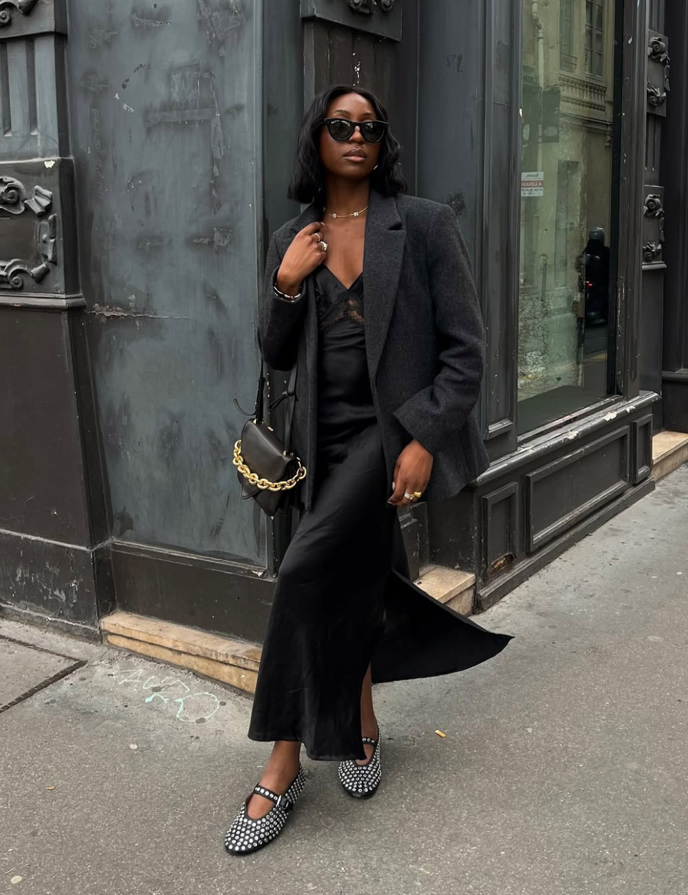 Um look bonito e fácil de reproduzir, destacando um vestido longo preto de tecido leve, complementado por um blazer cinza escuro. Os acessórios incluem óculos de sol elegantes e uma bolsa com corrente dourada.
