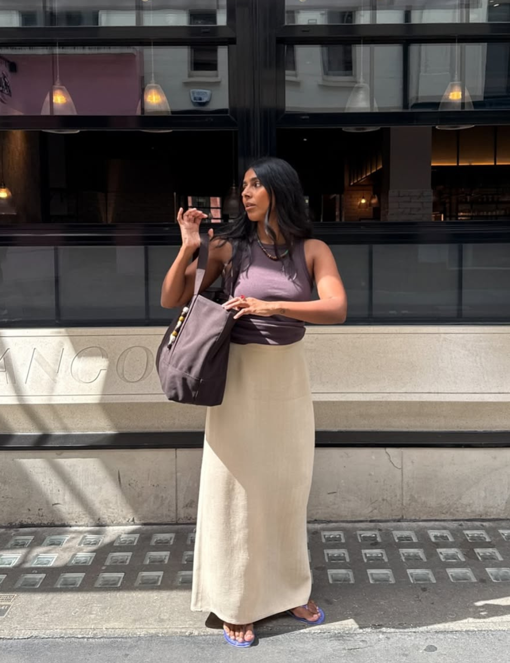 A imagem apresenta um look bonito e fácil, ideal para dias quentes. A pessoa usa uma blusa regata roxa e uma saia longa clara, que traz leveza e sofisticação. O acessório em destaque é uma bolsa marrom, prática e estilosa.