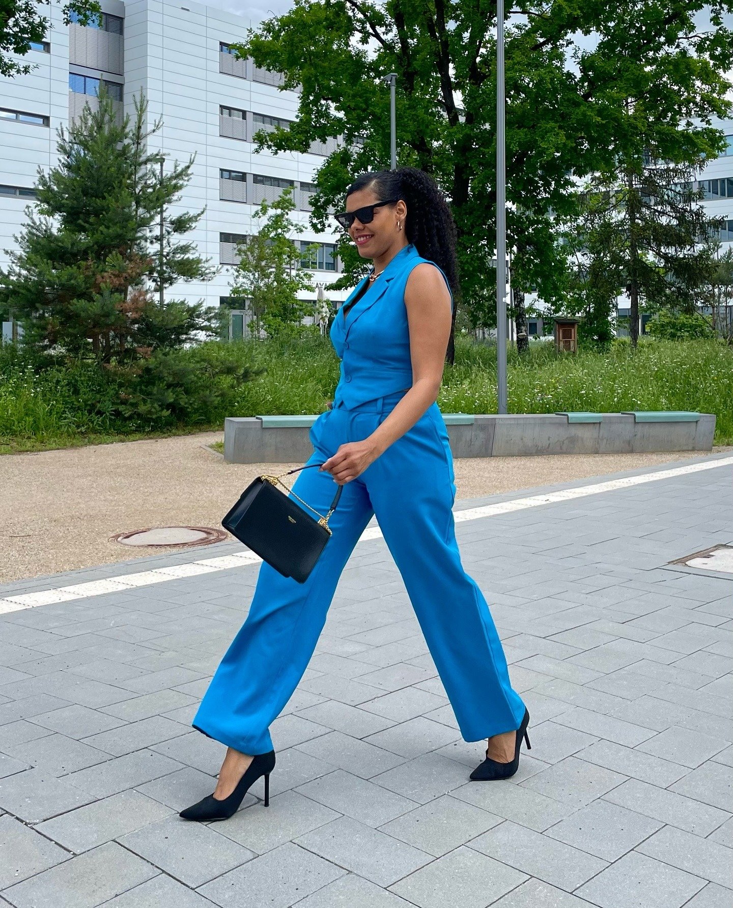 Look colorido para trabalhar: a modelo exibe um conjunto azul vibrante, que combina elegância e estilo. O traje, com calças largas e blusa ajustada, é complementado por sapatos de salto preto e uma bolsa de mão clássica. Óculos escuros adicionam um toque